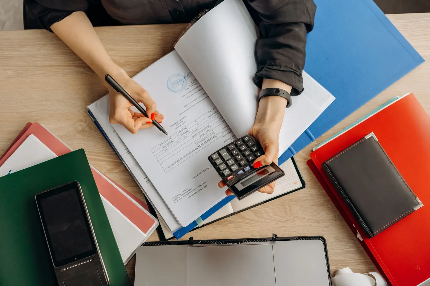 Hands hold a calculator and write in an open binder beside folders and a laptop.