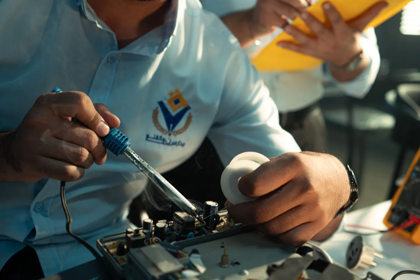 A technician solders components on an open circuit board while a coworker reviews notes on a yellow clipboard.