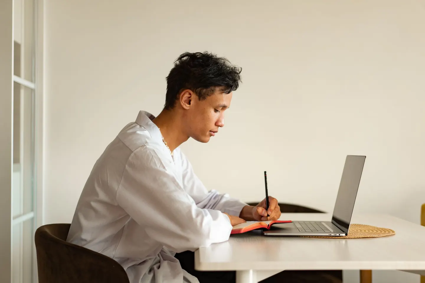 A man writing in a notebook beside his laptop at a bright desk.