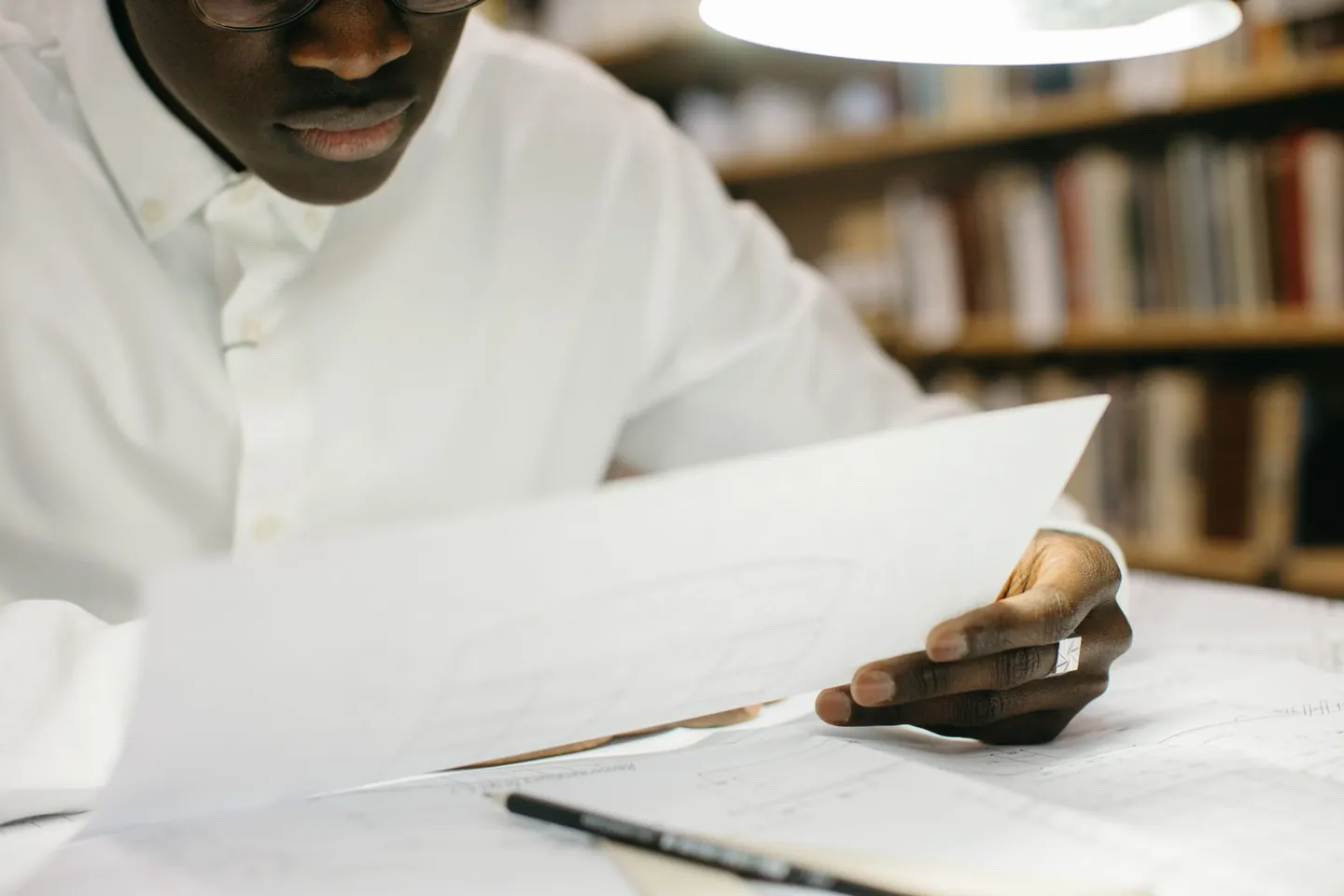 A man reviews papers at a library table beneath a desk lamp.