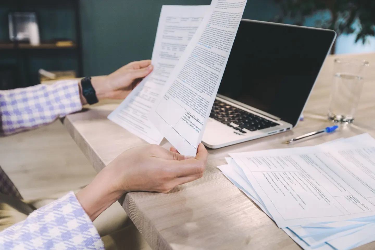 Two hands hold printed transcript pages above a desk with an open laptop and more papers beside it.