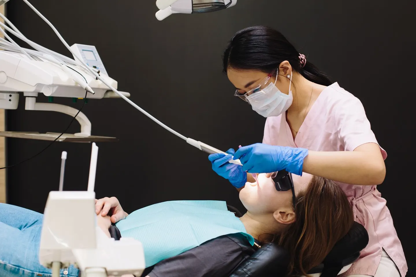 A dental hygienist in pink scrubs, mask and blue gloves cleans a reclining patient's teeth under an exam light.