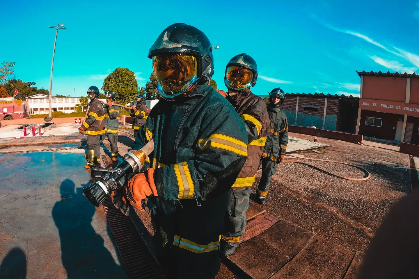Firefighters in helmets and reflective protective clothing handle a hose in an outdoor training area.