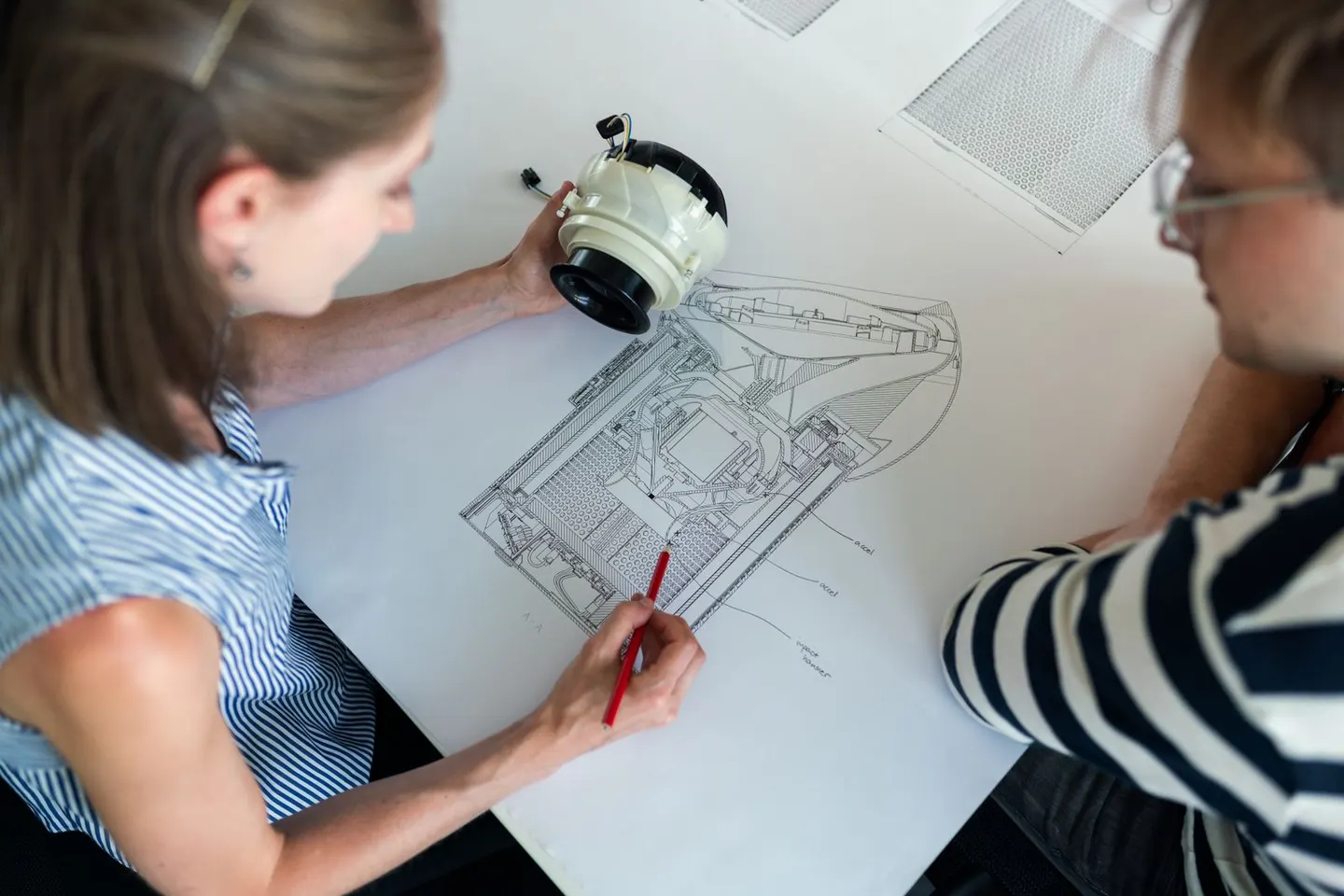Two people compare a mechanical component with a detailed drawing spread across a table.