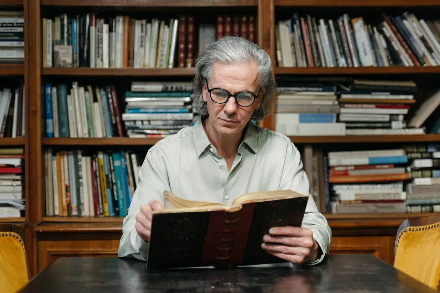 A gray-haired man wearing glasses reads an open book at a library table.