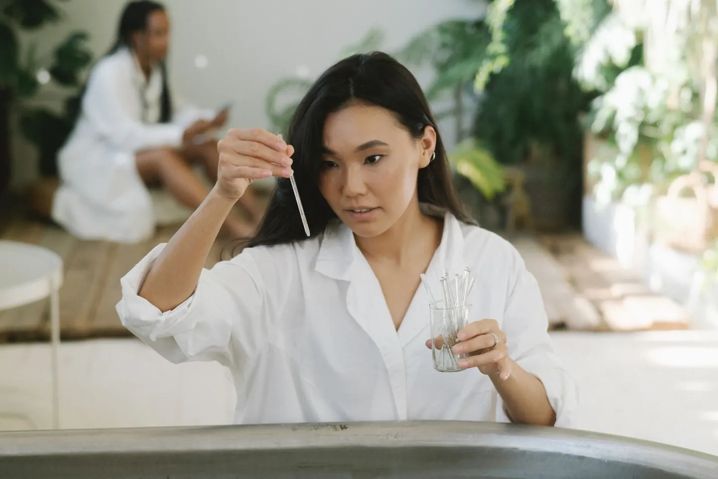 A woman examines a glass pipette beside a basin surrounded by plants.