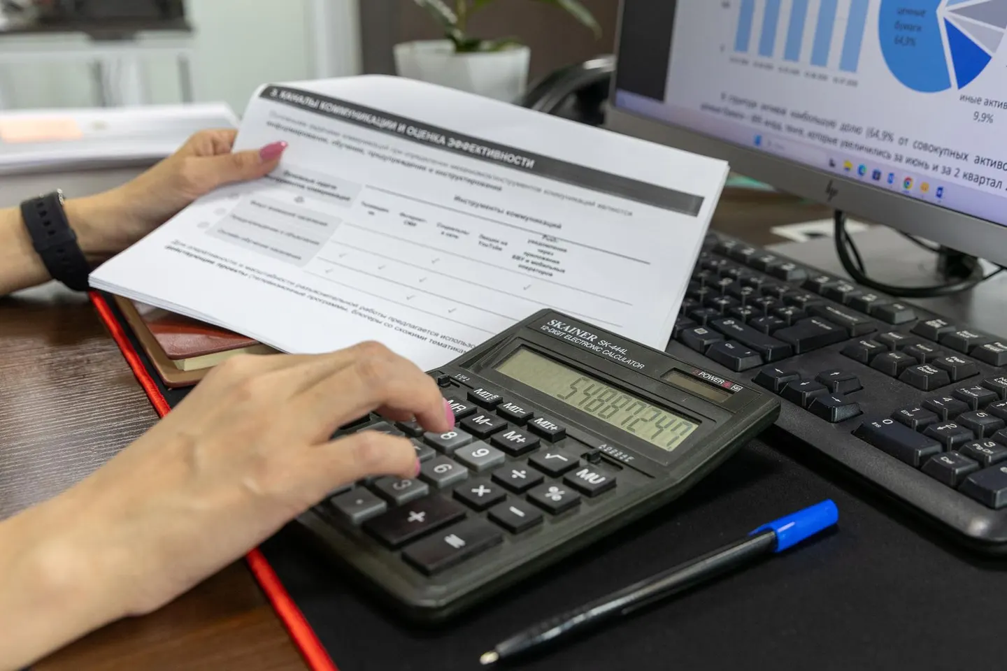 A person uses a calculator beside a financial document and a monitor displaying charts.