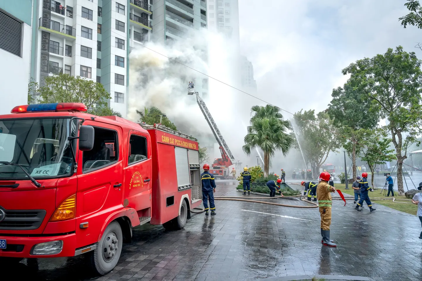 Municipal firefighters run hoses beneath an aerial ladder as smoke pours from a high-rise during a large training drill.
