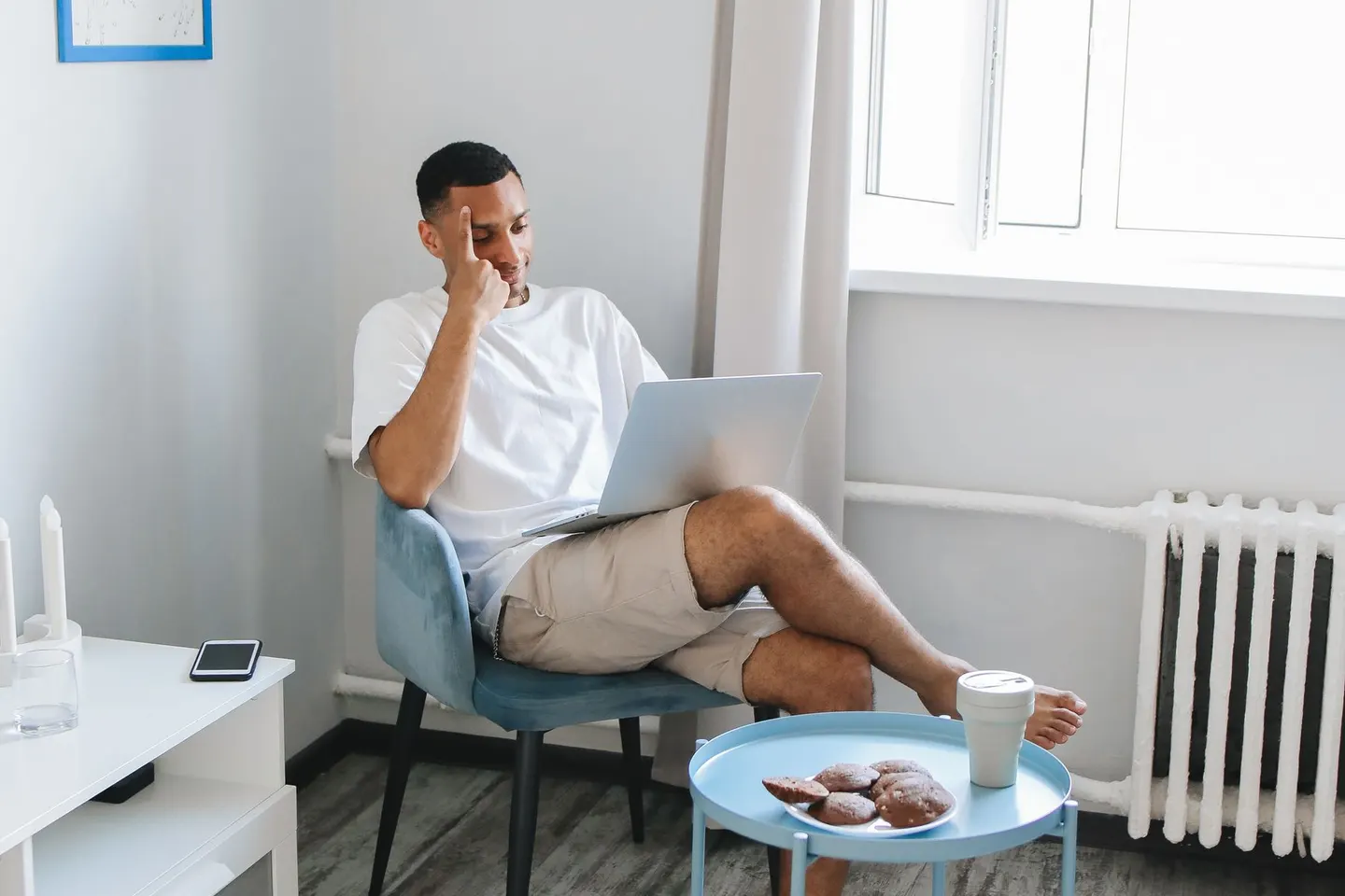 A man using a laptop in an armchair beside a sunlit window.