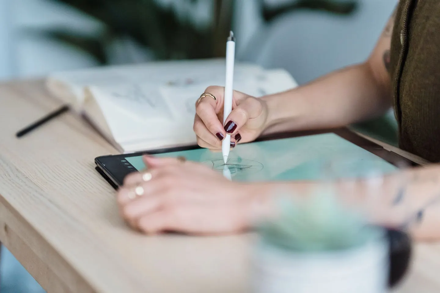 Hands use a white stylus on a drawing tablet resting on a wooden desk.