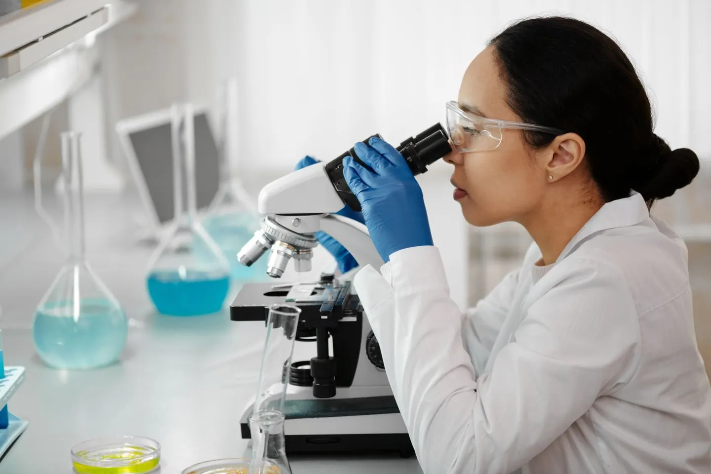 A woman wearing safety glasses and blue gloves looks through a microscope at a laboratory bench.