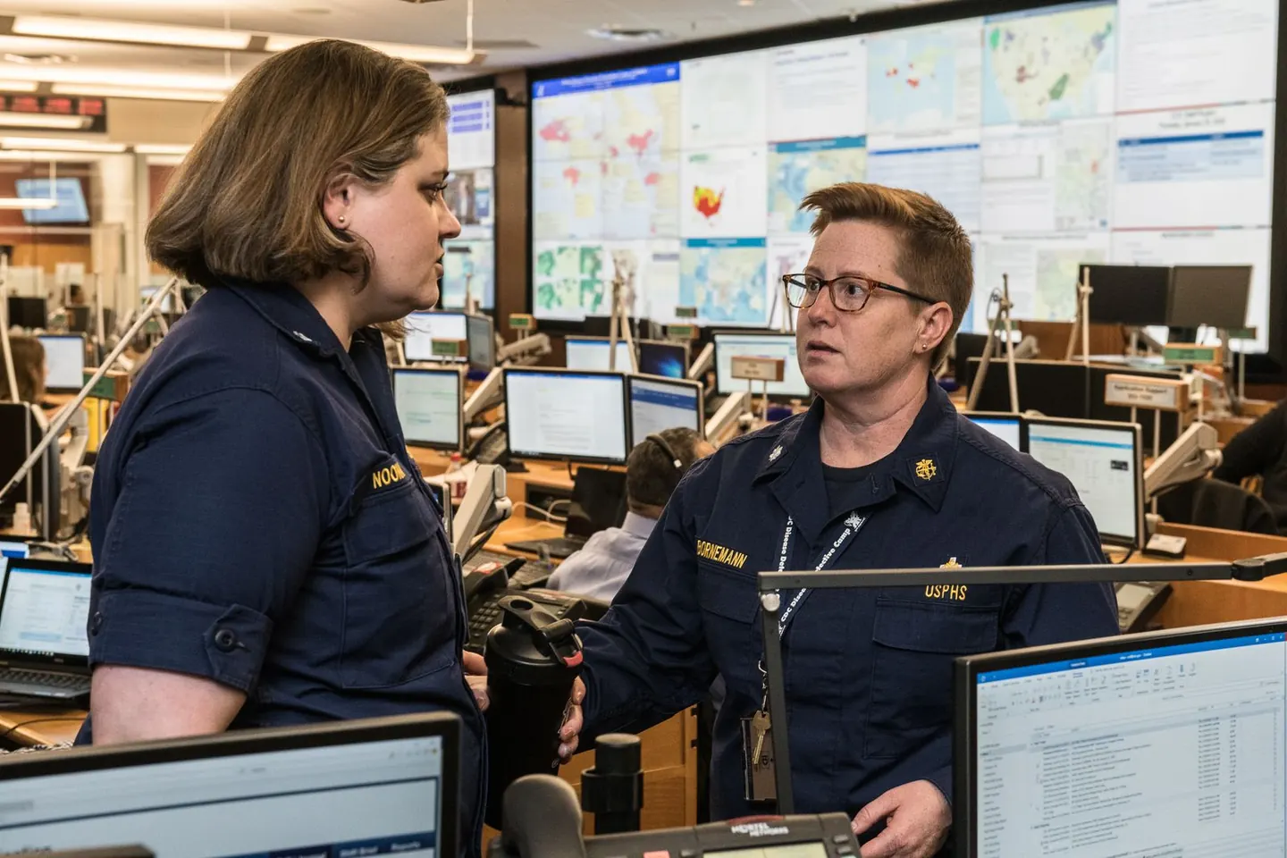 Two uniformed staff members talk in an operations center lined with computers and map displays.