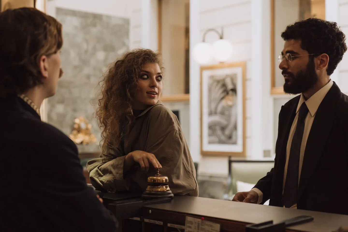 Three people talk across a reception desk with a service bell in the foreground.