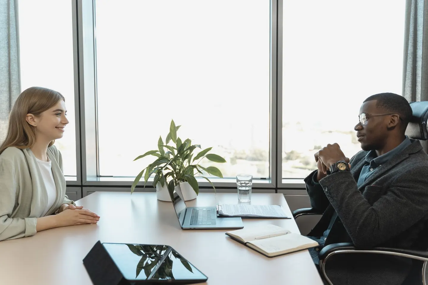Two people sit across an office desk with a laptop, tablet, and open notebook.