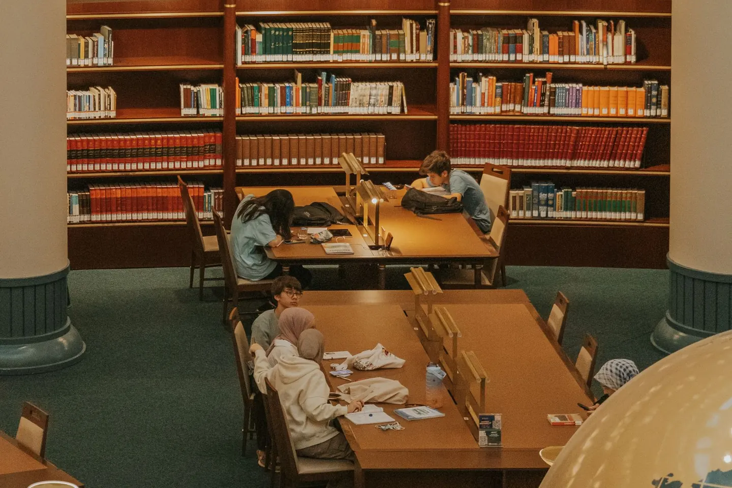 Adults read and work at wooden desks surrounded by library bookshelves.