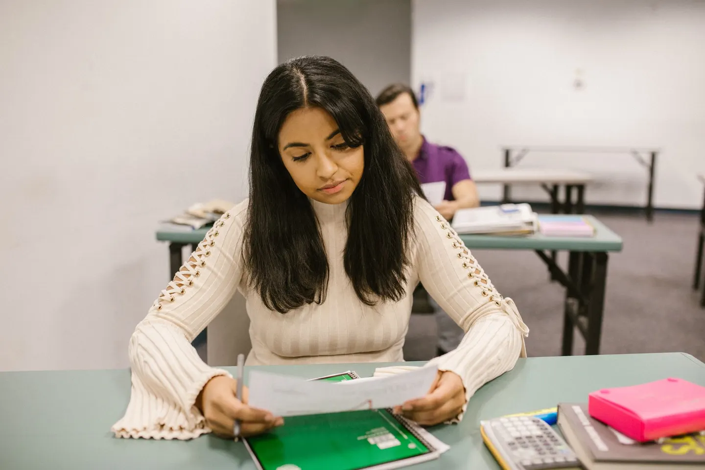 A college student at a classroom desk reads a graded paper beside a calculator and notebooks.