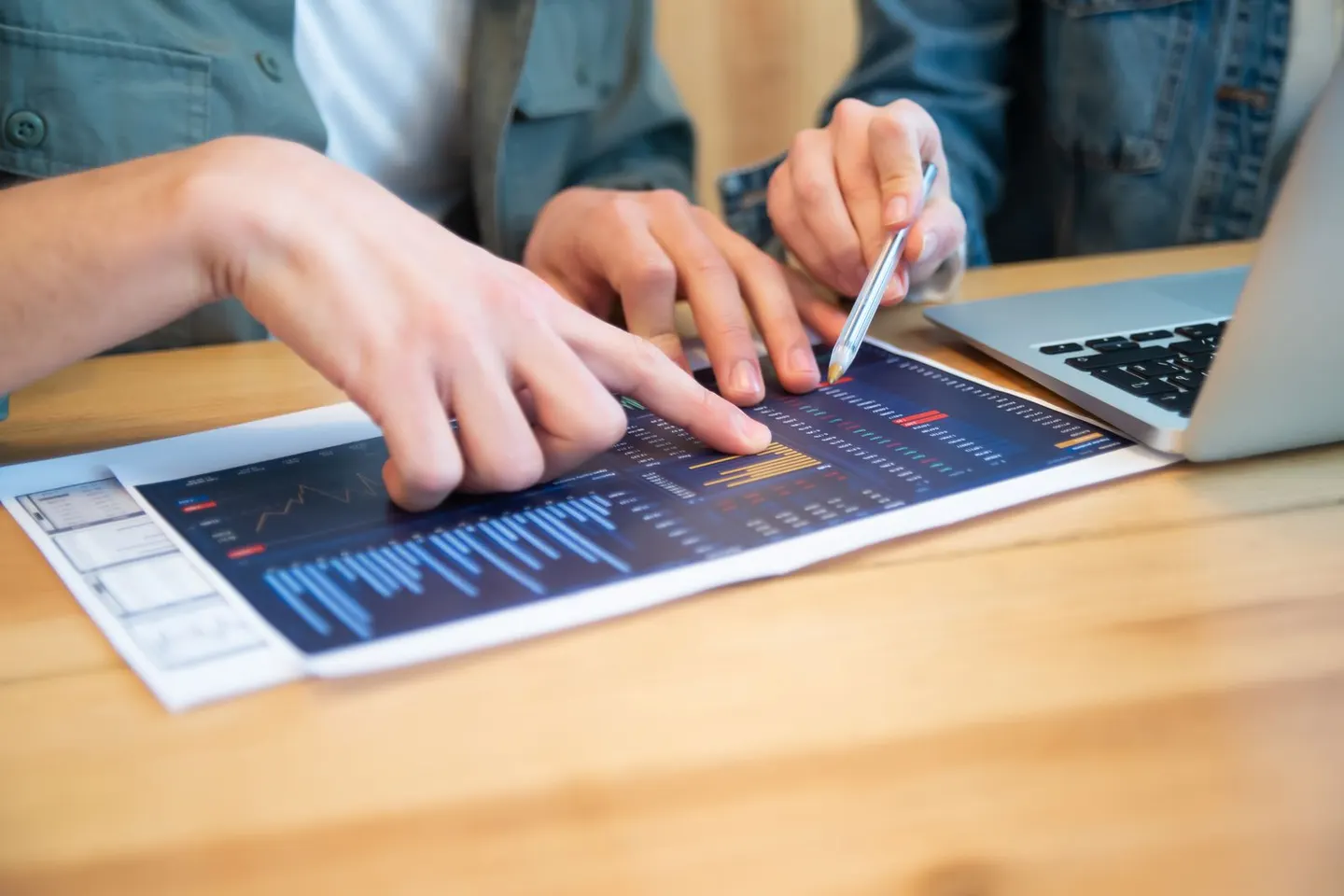 Hands point to printed charts beside a laptop on a wooden table.