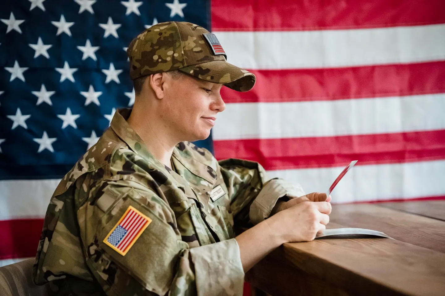 A service member in a camouflage uniform and cap reads a card at a wooden table in front of an American flag.