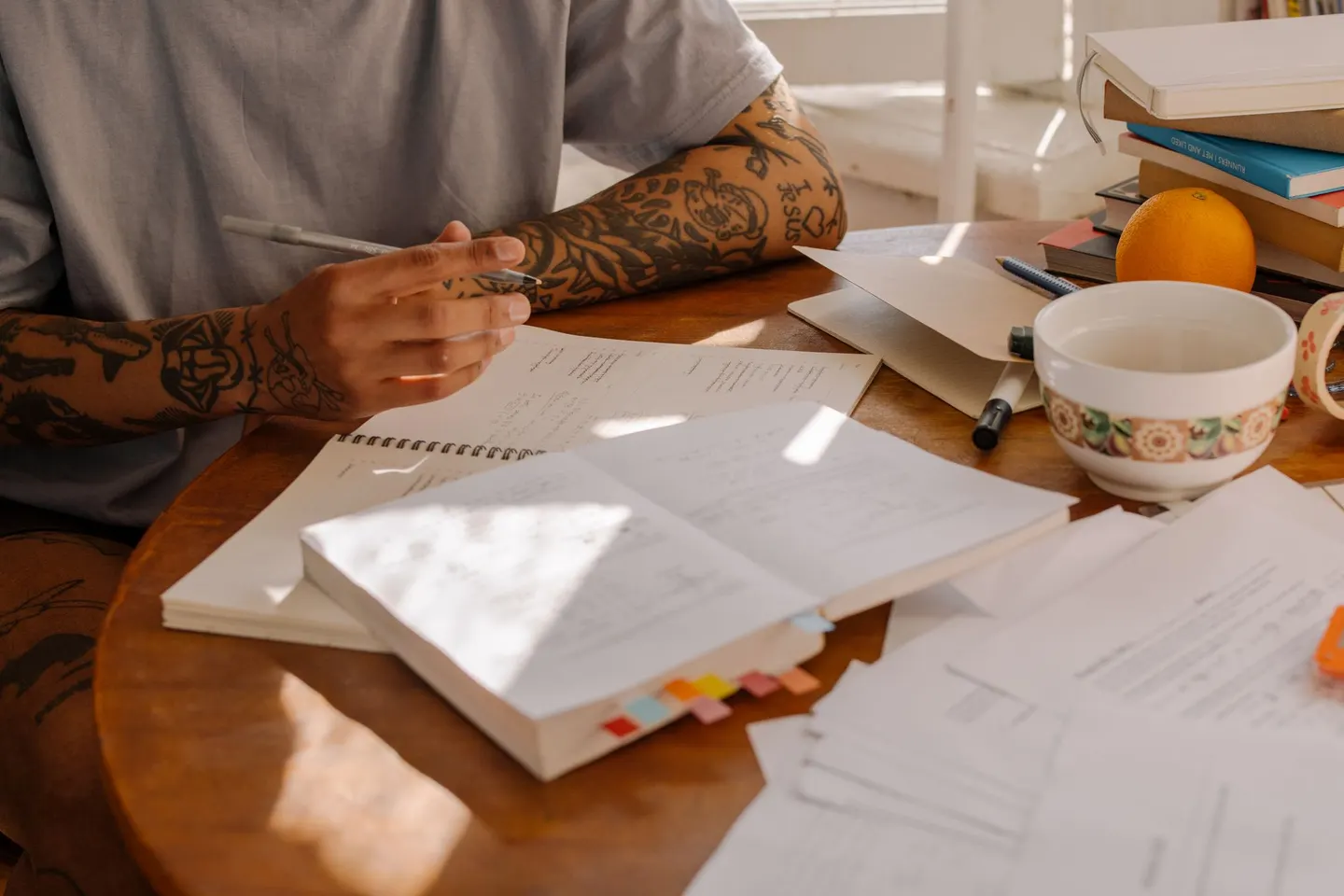 A student holding a pen works through notebooks, a tabbed book and loose papers spread across a round wooden table.