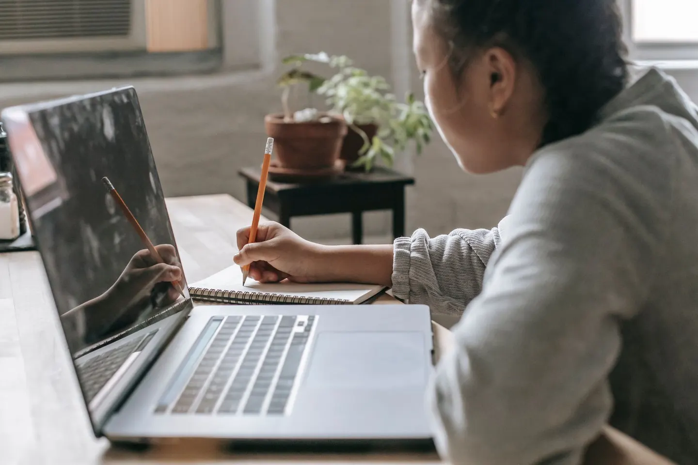 A woman writes in a notebook beside an open laptop at a table.