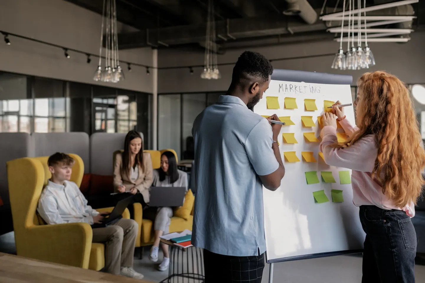 Two people arrange sticky notes on a flip chart while others sit nearby.