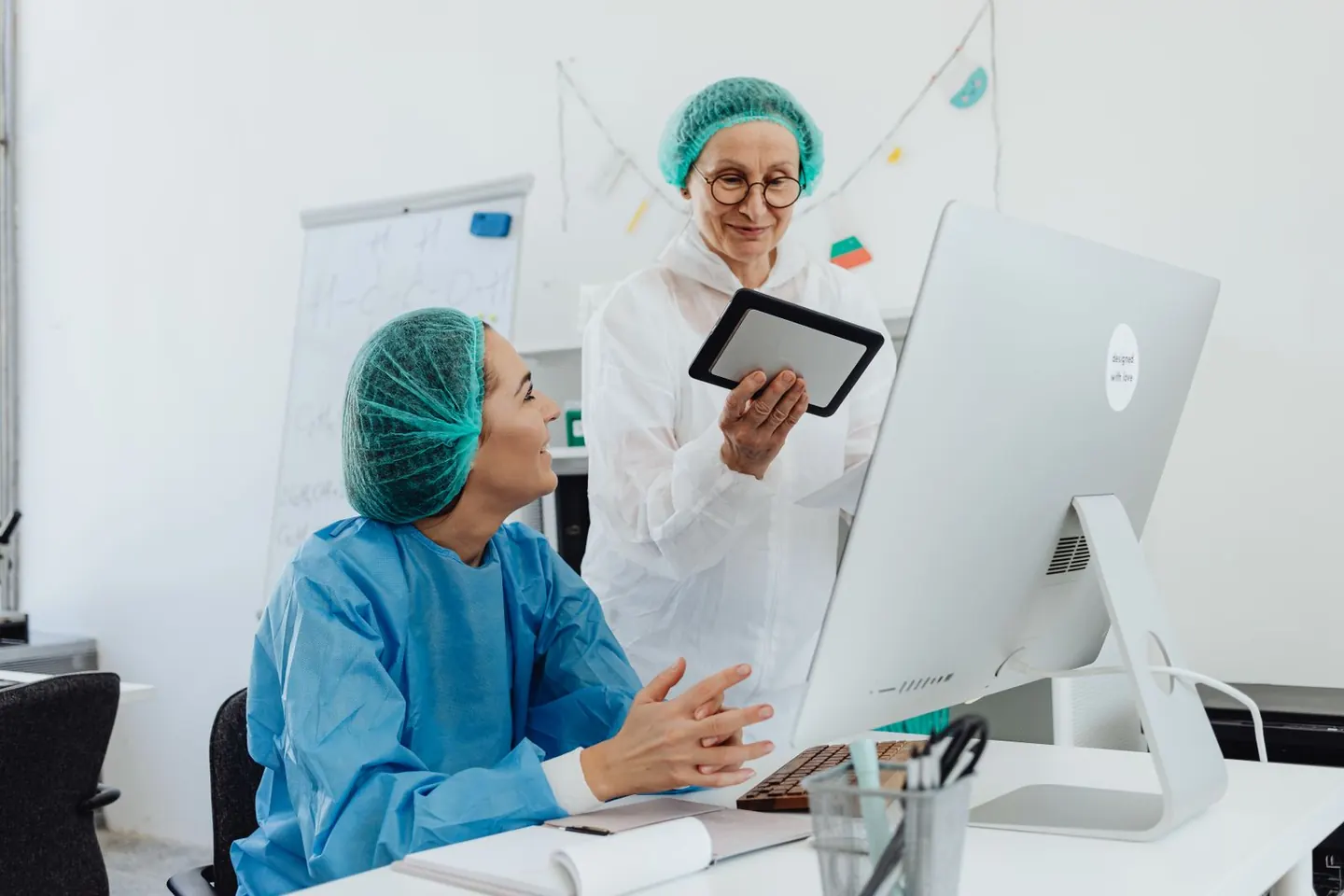 Two women in protective laboratory clothing discuss a tablet beside a desktop computer.