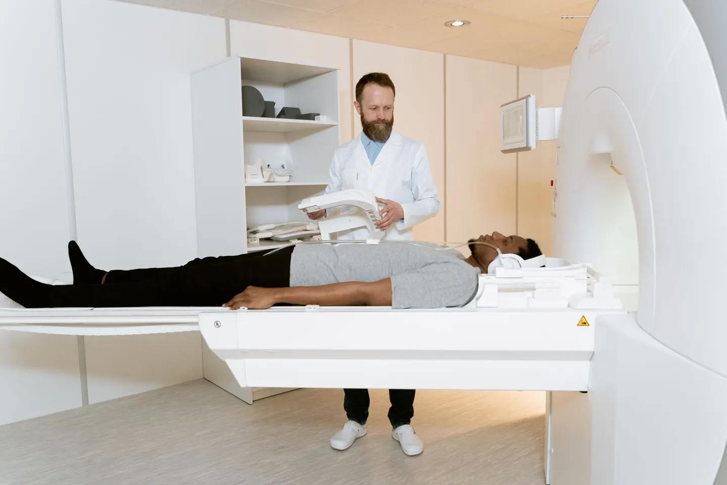 A radiologic technologist in a white coat positions an MRI coil while a patient lies on the scanner table