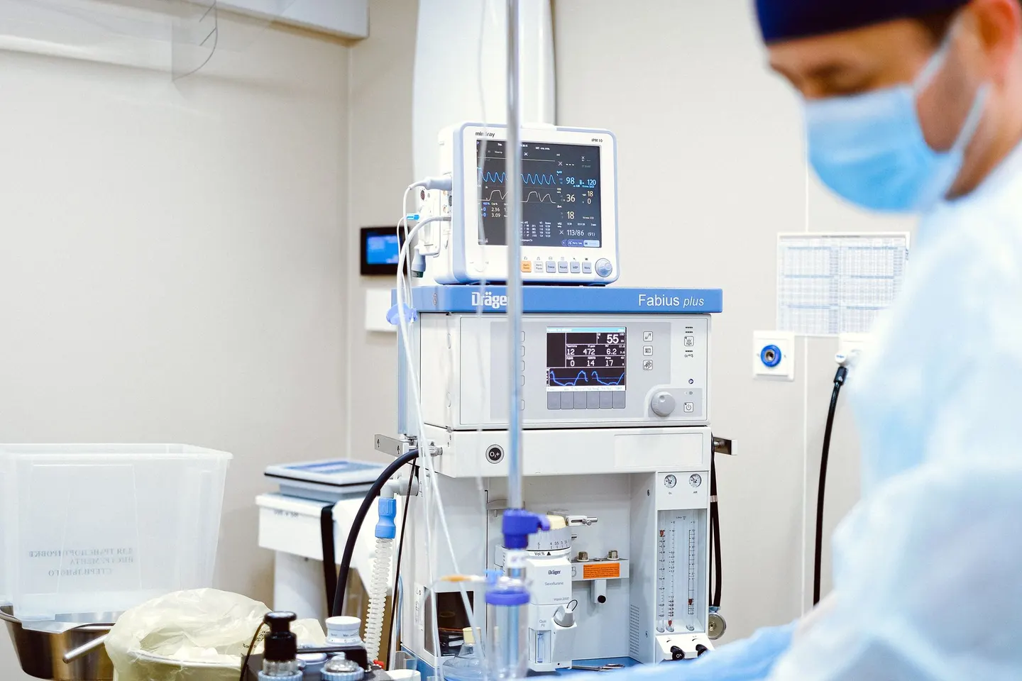 A clinician in a surgical cap and mask works beside a ventilator and patient monitor in a hospital room.