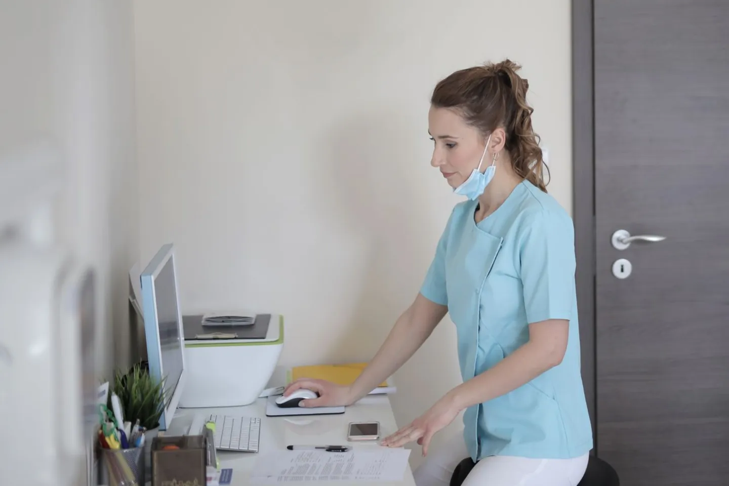 A woman in blue scrubs works at a desktop computer.