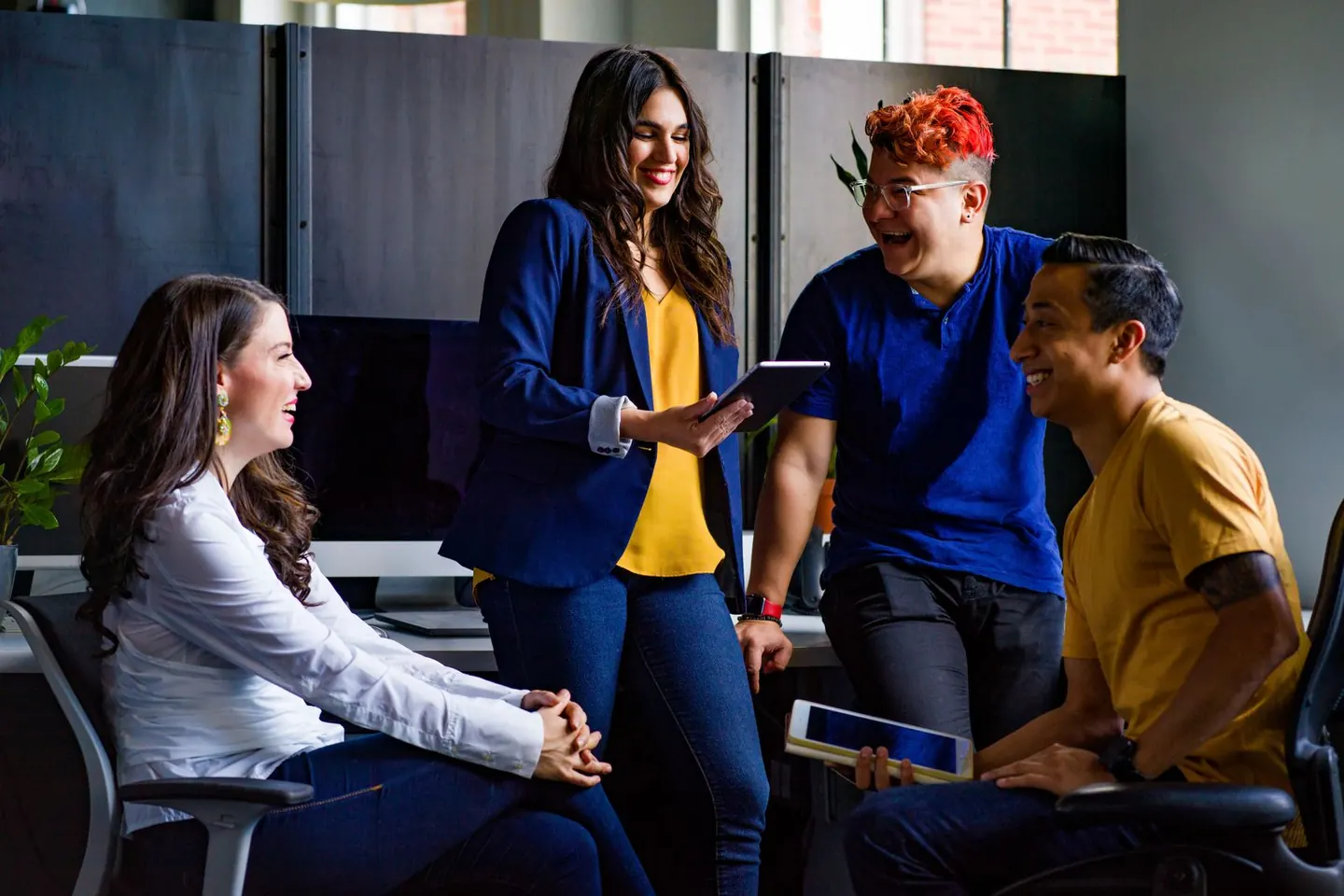 Four people gather around a tablet while talking in a bright shared workspace.