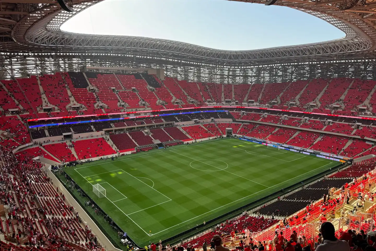 Spectators gather in red stadium stands around a soccer field beneath a sweeping roof.