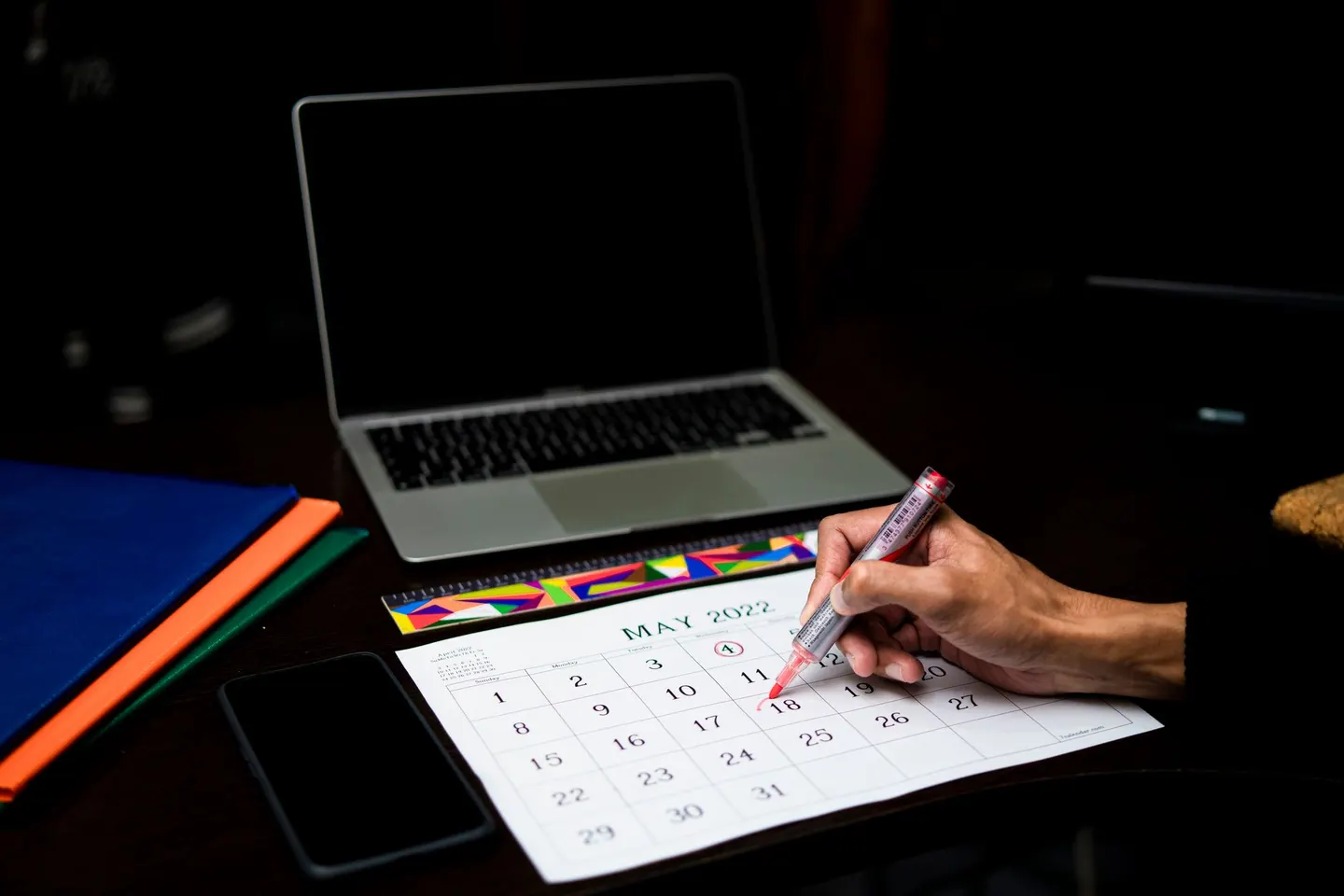 A hand circles a date on a printed monthly calendar in front of a laptop and phone.