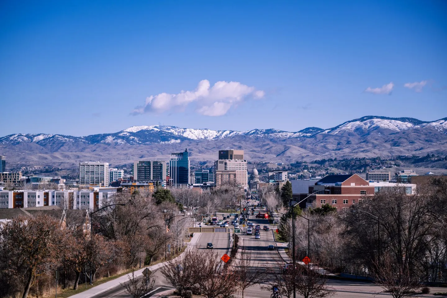 A tree-lined street leads toward downtown Boise with snow-dusted mountains behind it in Idaho.