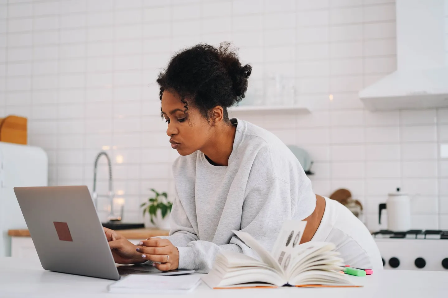 An adult student reads a course page on her laptop at a kitchen counter beside an open textbook.