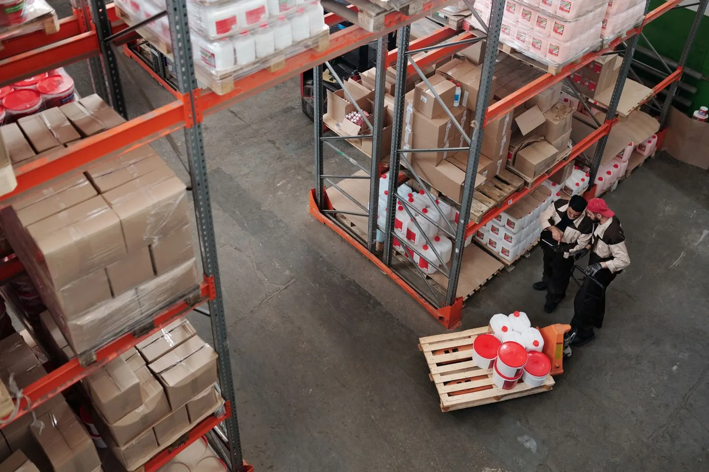 Two people stand beside loaded pallets between tall warehouse shelves viewed from above.