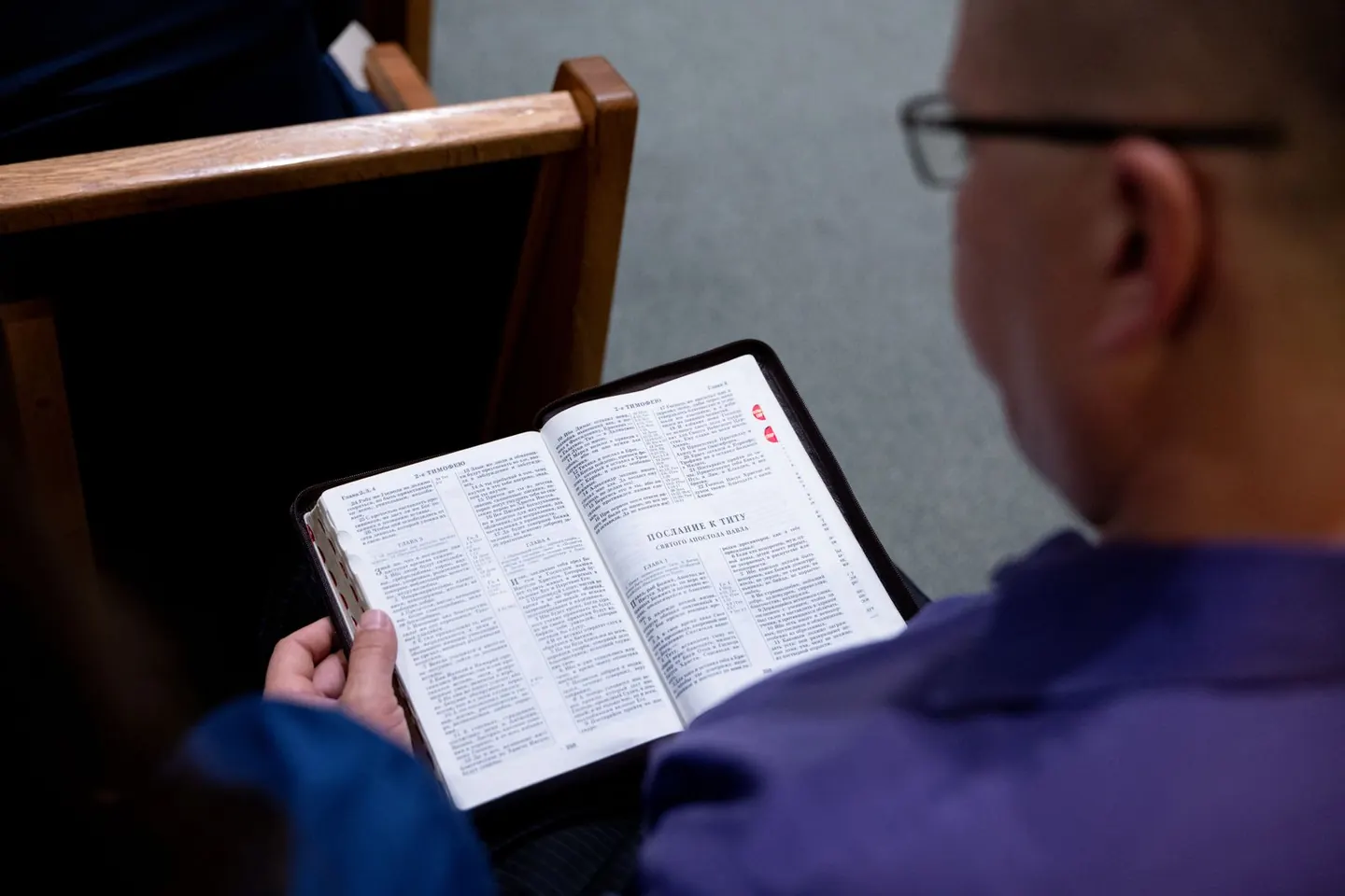 A seated man reads an open Bible beside rows of wooden church pews.
