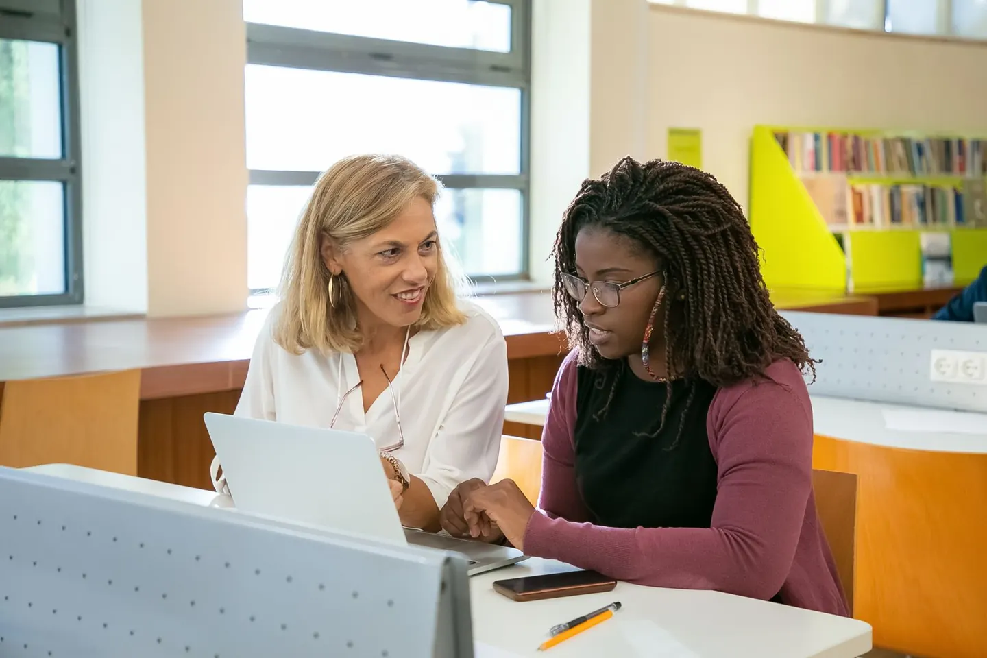 Two women discuss work on a laptop at a library study desk.